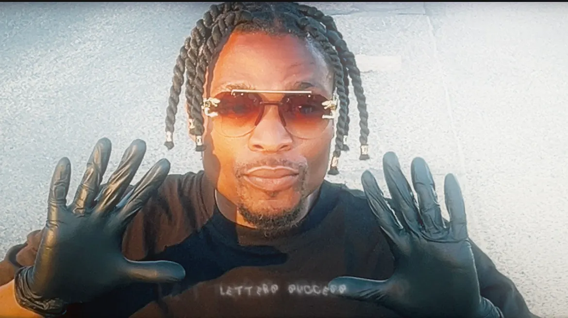 The barber, in braids and tinted glasses, holds up both black-gloved hands to the camera.