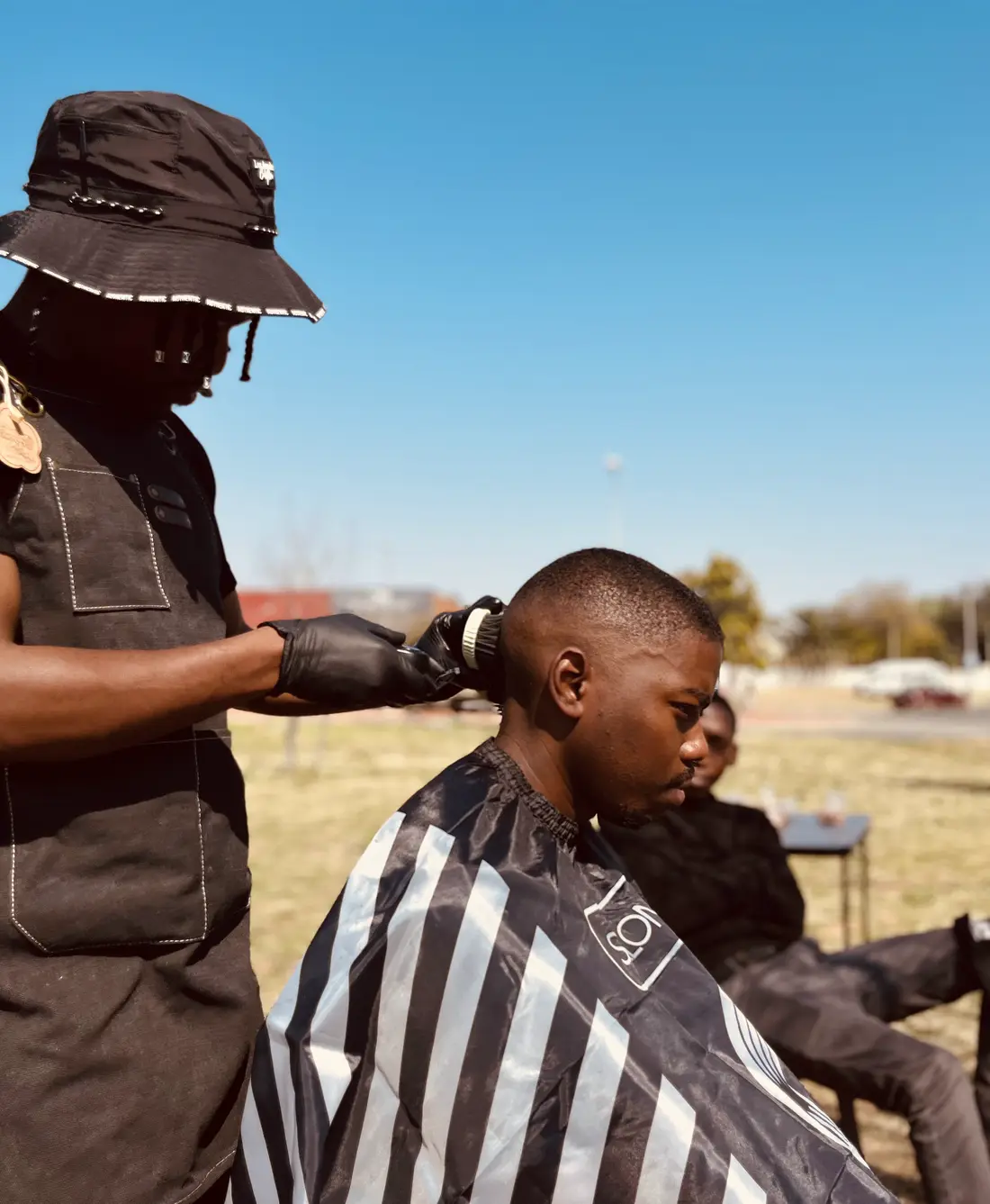 The barber, in a bucket hat and gloves, brushes a client's fresh cut in a field. Another man sits waiting on a chair behind them, with a road and open sky beyond.
