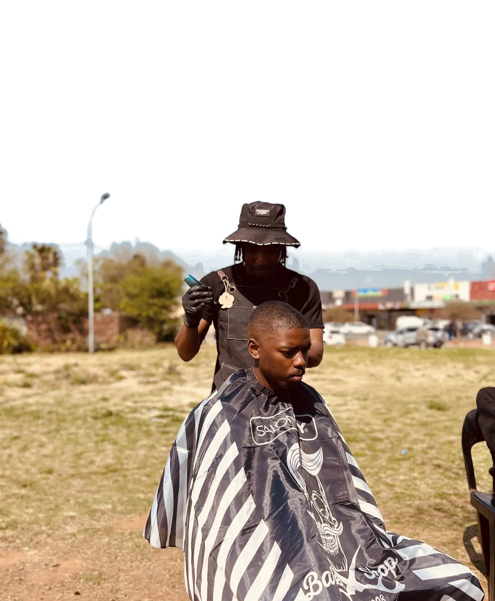 The KG Cutz barber, in a bucket hat and black gloves, works on a client in a striped cape in an open field under a clear blue sky.